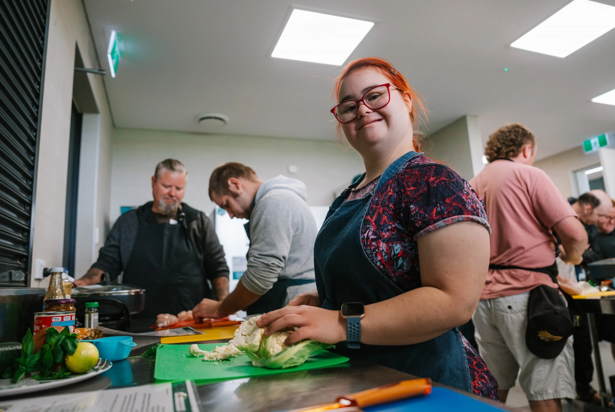 A young adult with disability wearing an apron chops cauliflower during a group cooking activity, with other participants preparing food in the background.